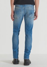 Uske plavi traperice slim-fit kroja s izblijedjelim pranjem, pet džepova i šavovima na zadnjem dijelu. Nosi crne čizme.