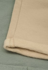 Beiger Pulloverbund mit einer weichen, flauschigen Textur, dargestellt neben einem grünen Stoff. Sichtbare Nähte entlang der Kanten des Bunds.