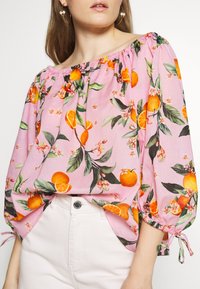 Donna che indossa una blusa rosa a spalle scoperte con stampa di frutti arancioni e fiori, abbinata a pantaloni bianchi a vita alta e orecchini a cerchio dorati.