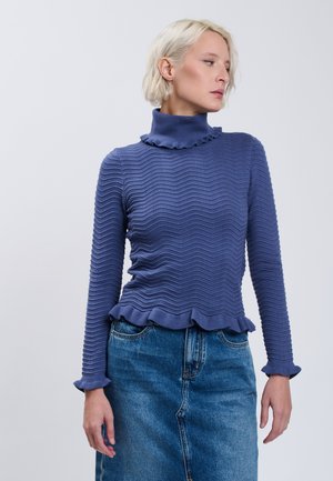 Kvinde med kort blondt hår iført en blå tekstureret rullekravetrøje og en blå denimnederdel, der kigger til siden mod en ensfarvet baggrund.