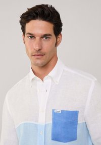 Camicia bianca in lino con tasca blu e design a blocchi di colore. La camicia presenta un colletto con bottoni e una vestibilità casual.
