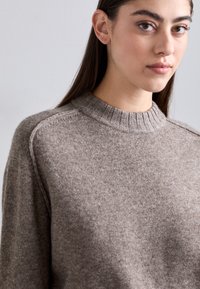 Nainen, jolla on pitkät ruskeat hiukset, pukeutuneena vaaleanruskeaan villapaitaan, jossa on ribbireunus kauluksessa, yksinkertaista taustaa vasten.