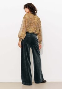 Blouse légère et à motifs dans des tons dorés et violets avec des manches longues ; pantalon en velours côtelé large et sombre ; arrière-plan minimaliste.