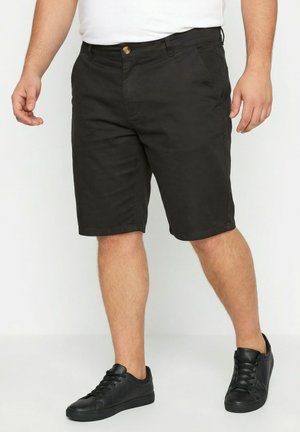 Mand iført sorte knælange shorts, sorte sneakers og hvid skjorte, stående på en ensfarvet hvid baggrund.