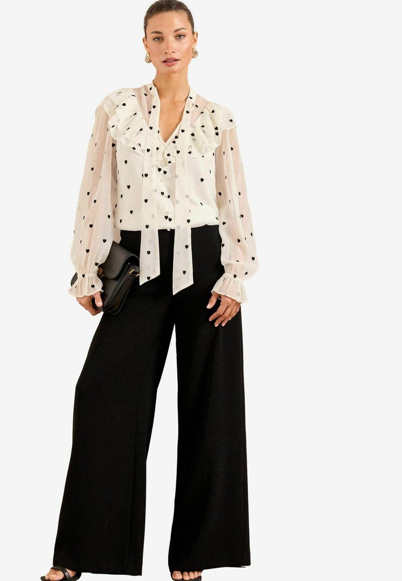 Sheer white blouse with black heart patterns and ruffled neckline, paired with wide-leg black trousers. Accessories include a black clutch.