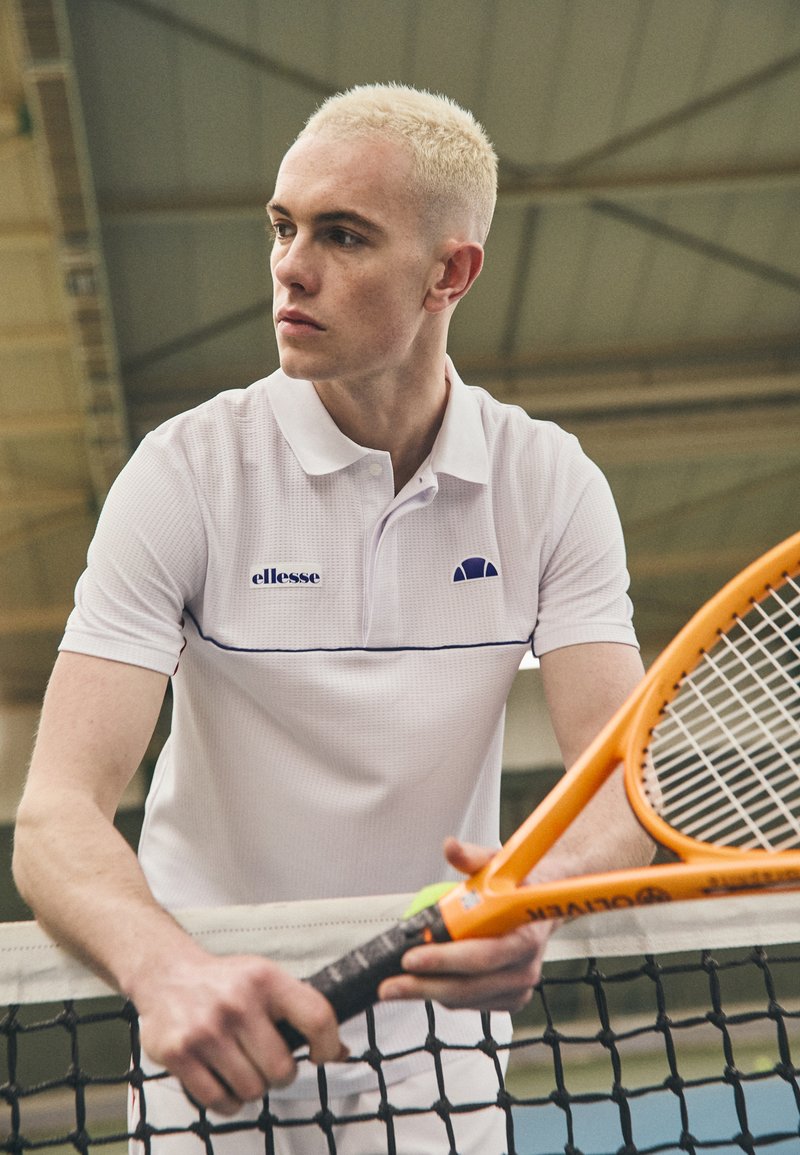 Hvit polo skjorte med teksturfinish, blå aksentstripe og logo. Spilleren holder en oransje tennisracket ved et nett i en innendørs sportsanlegg.
