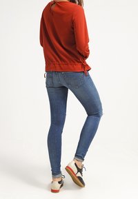 Haut à manches longues couleur rouille avec un trou clé au dos, associé à un jean en denim bleu slim et des chaussures à lacets multicolores. Textures lisses dans l'ensemble.