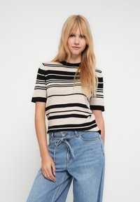 Jeune femme aux longs cheveux blonds portant un haut à manches courtes beige et noir à rayures et un jean bleu clair avec ceinture à nouer, posant devant un fond blanc.