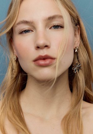 Nærbillede af en ung kvinde med langt lyst hår, der bærer hængende guldøreringe formet som blomster mod en blå baggrund.