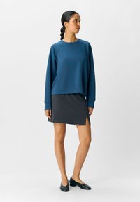 Blauer Baumwoll-Sweatshirt mit geripptem Textur, kombiniert mit einem dunkelgrauen Rock mit seitlichen Schlitzen. Schwarze flache Schuhe vervollständigen das Outfit.