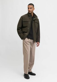 Férfi sötét olajzöld dzsekiben, fekete ingben, bézs nadrágban és fekete cipőben áll, az egyik kezét a zsebébe dugva, fehér háttér előtt.