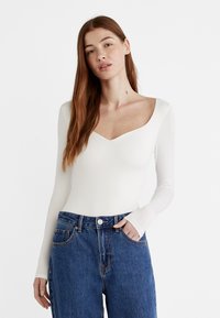 Hvid langærmet bodystocking med dyb V-udskæring, parret med højtaljede blå denimjeans. Glat tekstur, tætsiddende design og minimal detaljering.