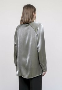Chemise à manches longues en tissu vert olive métallisé, avec une texture brillante, une coupe loose, un ourlet arrondi et des poignets à boutons.