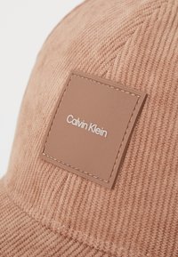 Pruunist korduroüst valmistatud müts, millel on ruudukujuline nahast patch, kus on "Calvin Klein" logo. Tekstuuritud kangas ja õmblustooted tõstavad disaini esile.