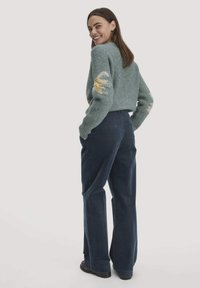 Lichtgroene gebreide trui met gele en witte accenten, gecombineerd met donkerblauwe corduroy broek, gezien van achter.