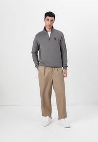 Felpa grigia con cerniera e logo sul petto, abbinata a pantaloni beige a gamba larga e sneaker bianche, su uno sfondo semplice.