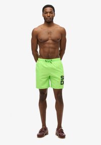 Costumi da bagno verde neon con una stampa grafica nera, vita con laccetto e un tessuto liscio e leggero. Indossati con scarpe marroni.