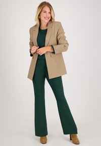 Beige blazer over een donkergroene top en bijpassende flared pants. Bruine hakken. Glad materiaal met gestructureerde tailoring en subtiele manchetdetails.