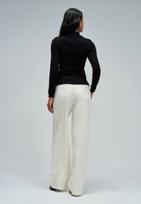 Woman with long dark hair wearing black long-sleeve top, white wide-leg pants, and black heels, standing facing a plain light gray background.