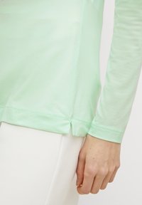 Jasnozielona koszula z długim rękawem wykonana z miękkiego materiału, z zaokrąglonym dołem i bocznymi rozcięciami. Gładka faktura i elastyczny materiał.