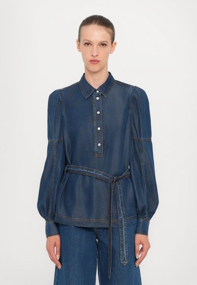 Chemisier en denim avec un col, une fermeture à boutons, et des manches bouffantes. Il présente une taille ceinturée et des coutures bleues sur un tissu bleu foncé.