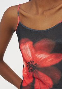 Femme portant un haut à fines bretelles foncé avec un grand imprimé de fleurs rouges, épaule, clavicule et haut de la poitrine visibles.