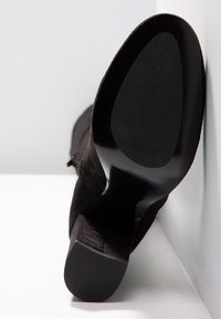 Botte noire en daim à talons hauts vue de la semelle, reposant contre un mur blanc et un sol blanc.