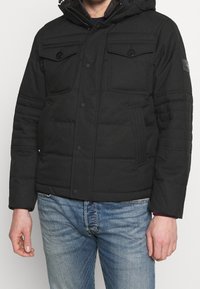 Mann in einer schwarzen gepolsterten Jacke mit geknöpften Brusttaschen und blauen Jeans, der vor einem schlichten hellen Hintergrund steht.