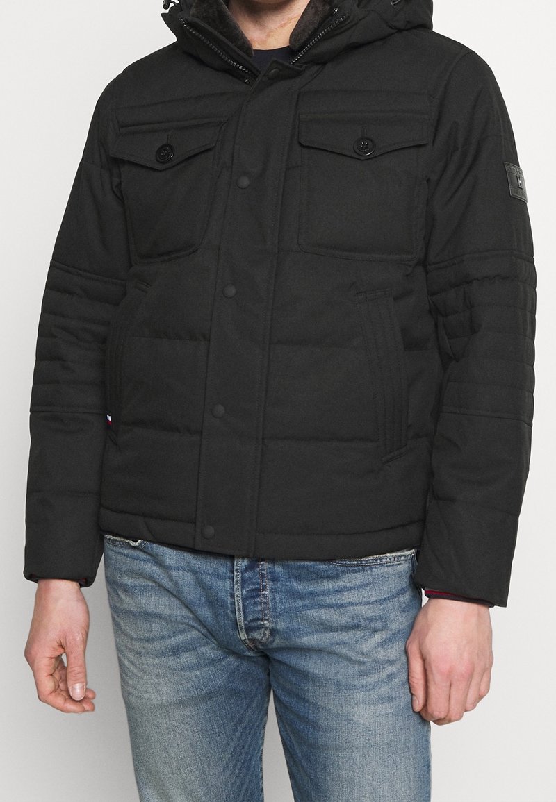 Mann in einer schwarzen gepolsterten Jacke mit geknöpften Brusttaschen und blauen Jeans, der vor einem schlichten hellen Hintergrund steht.