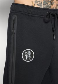 Sorte joggers med elastisk talje, sidelommer med lynlås og et hvidt broderet cirkulært logo, der viser en løve og initialerne "CFC."