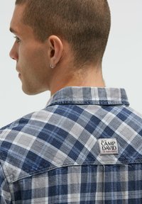 Blau- und grau kariertes Hemd mit einer Denim-Textur, versehen mit einem Etikett am Kragen und kontrastierenden Karomustern. Kurzärmel sind sichtbar.