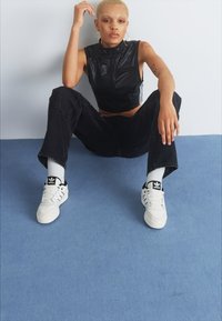 Fekete, rövidített trikó, laza fekete farmernadrág, fehér sneakers fekete részletekkel és fehér zokni; a személy egy kék padlón ül.