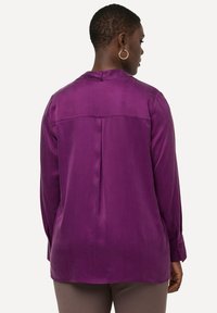 Paarse blouse met lange mouwen, met een gladde textuur en een plooi detail aan de achterkant. Beschikt over knoopmanchetten en een ronde halslijn.