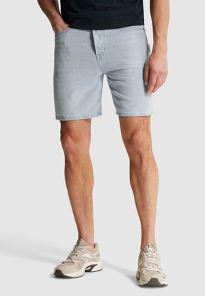 Muž nosi svetlomodré džínsy kraťasy, béžové tenisky a čiernu košeľu, stojí s jednou rukou v vrecku na sivom pozadí.