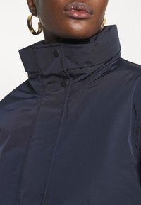 Närbild av en person som bär en marinblå jacka med hög krage, svarta tryckknappar och stora guldörhängen.