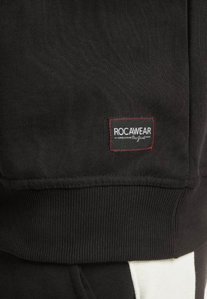Juodas sportinis džemperis su iškilnia tekstūra, su raudonu ir baltu Rocawear logotipo užkliju, o apatiniame skyriuje kontrastingos baltos detalės.