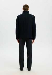 Cappotto in lana scuro con colletto alto, progettato con una silhouette strutturata. Abbinato a pantaloni texturizzati blu navy e scarpe nere.