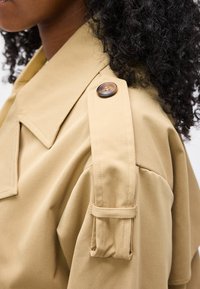 Κοντινό πλάνο λεπτομέρειας στον ώμο ενός μπεζ παλτού τύπου trench coat με κουμπωμένη επιγονατίδα, που φοριέται από πρόσωπο με σγουρά μαλλιά.