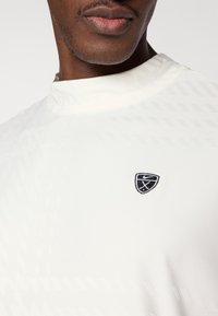 Biała sportowa koszulka z teksturowanymi ukośnymi wzorami, wysokim kołnierzem i małym czarnym haftowanym logo na piersi.