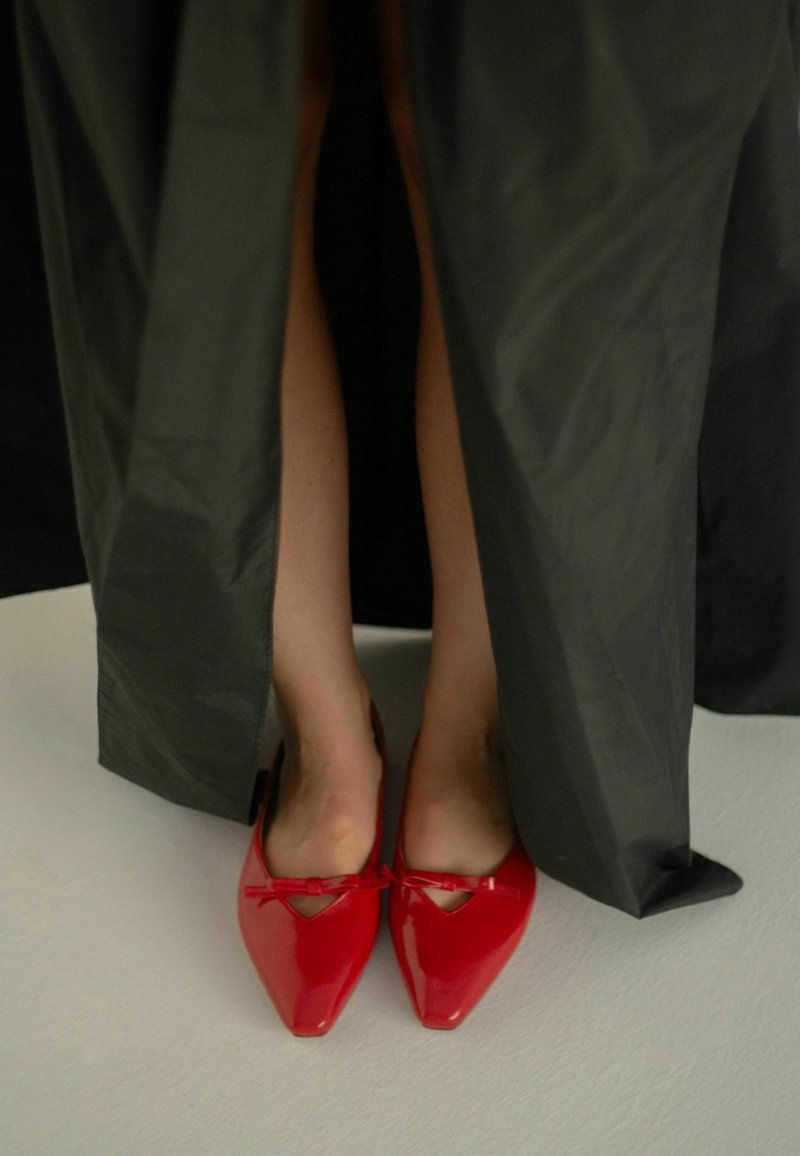 Scarpe in vernice rossa con punte appuntite e piccoli fiocchi sulla parte anteriore, posizionate sotto un abito nero fluente.