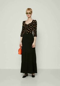 Μαύρη φλοράλ μπλούζα με φουσκωτά μανίκια, συνδυασμένη με μακριά μαύρη φούστα. Αξεσουάρ περιλαμβάνουν μια μικρή κόκκινη τσάντα χεριού και μαύρα φλατ παπούτσια.