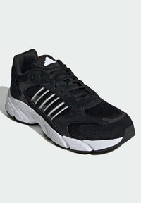 Svart atletisk sneakersko med övre del i mesh och mocka. Har tre silverränder och en vit sula med texturerade mönster.