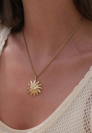 Goldene, sonnenförmige Anhänger an einer verdrehten Kettenhalskette, getragen am Hals einer Frau, mit einem cremefarbenen, gestrickten ärmellosen Oberteil sichtbar.