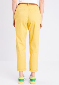 Pantalons jaunes à coupe slim, avec des ourlets retroussés et des poches arrière. Associés à des baskets vertes et blanches et une ceinture tressée marron.