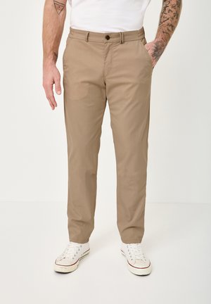 Man wearing beige straight-fit trousers, white sneakers, and a white shirt, standing with one hand in pocket, tattooed arms visible.