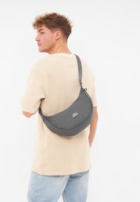 Borsa a spalla grigia e morbida, dalla forma curva, con etichetta frontale e tracolla regolabile, indossata sopra una camicia chiara.