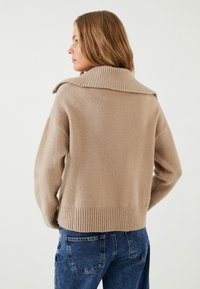 Beige strikket sweater med en bred ribbet krave, der har dropped skuldre og en ribbet kant. Blød tekstur, afslappet pasform, parret med blå jeans.