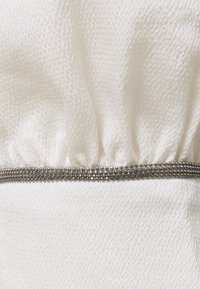 Vit tyg med en texturerad, plisserad sektion och en silverpärlband längs botten. Det övergripande utseendet är slätt och fint.