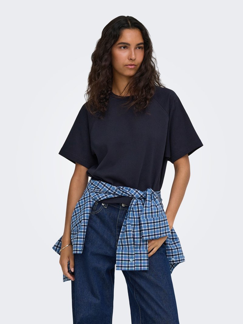 Femme aux longs cheveux bouclés portant un t-shirt noir à manches courtes, un jean bleu et une chemise à carreaux bleue nouée autour de la taille.