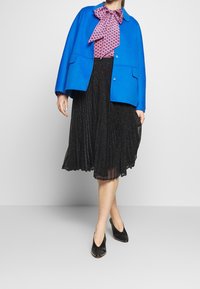 Chaqueta de lana azul brillante con dos bolsillos, blusa estampada en rosa, falda negra plisada con brillo y zapatos negros con acentos de tachuelas.